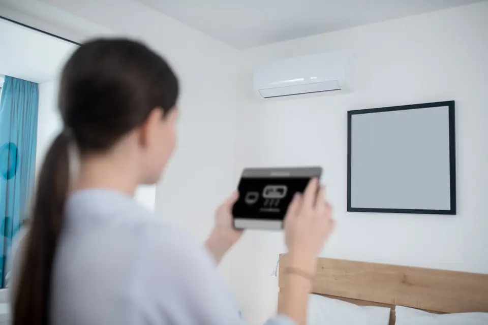 Frau steuert das Innengerät einer Klimaanlage im Schlafzimmer mit einem Tablet, von hinten fotografiert